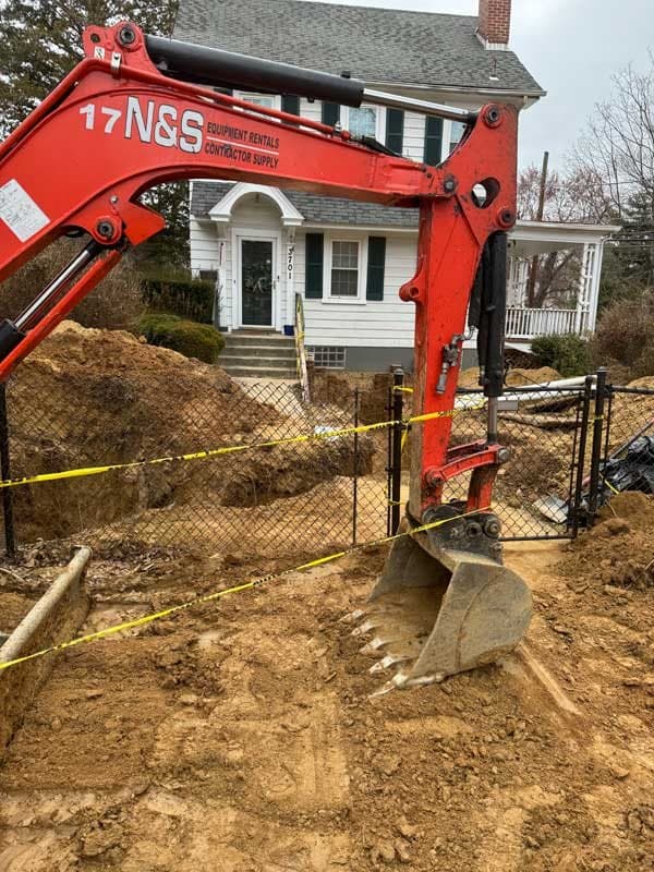 Red mini excavator digging a small access hole in the fenced front yard of a white house, with caution tape around the work area