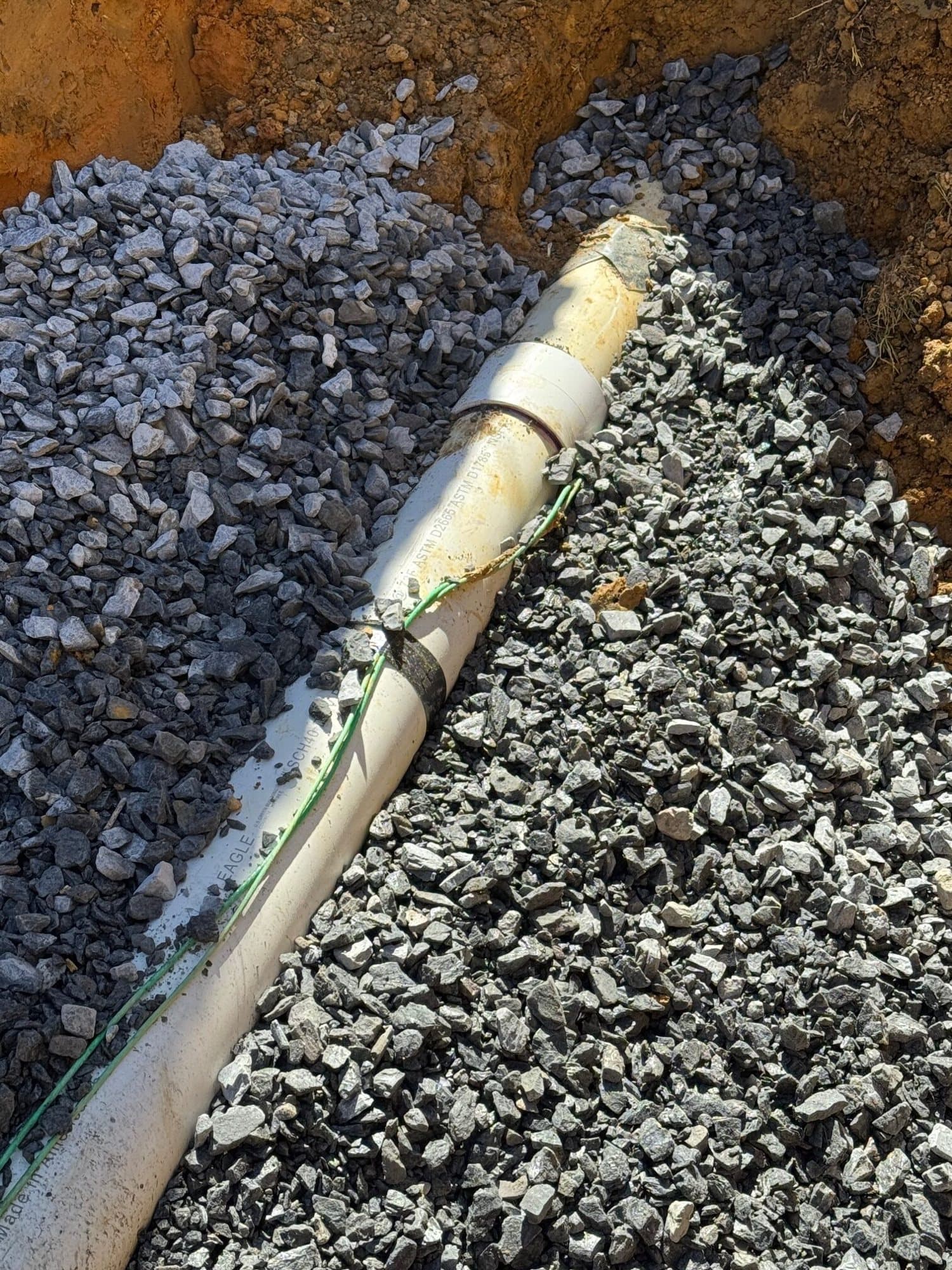 New white PVC sewer lateral with a coupling, bedded in crushed stone with green tracer wire taped along it